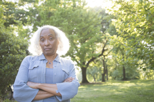 A senior woman with gray and white hair crosses her arms and looks disparagingly at the camera. 