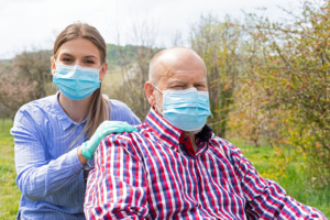 Elderly disabled man with mask sitting in wheelchair, assisted by young female caregiver outdoors