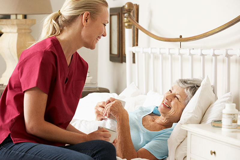 A caregiver brings a cup of tea to an older woman in bed