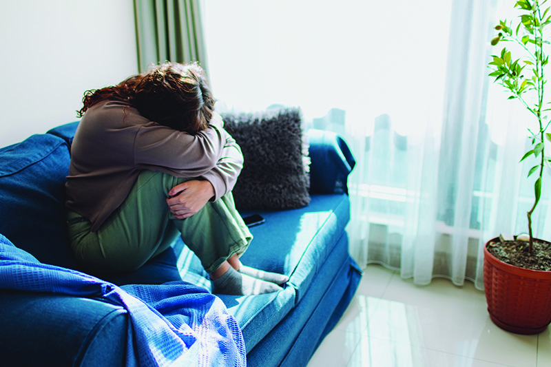 A female family caregiver sits on couch with knees pulled up and head buried in her arms on top of her knees.