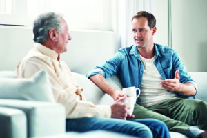 Adult son and elderly father sit on a couch having a conversation.