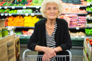 An older woman stands in the grocery store, leaning on her cart with a calm expression on her face.