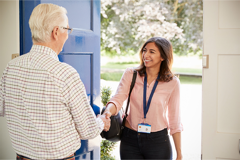 An older man is standing by the front door, shaking hands with a new caregiver as he welcomes her into his home.