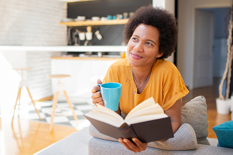 A woman who knows how caregivers can prioritize privacy smiles as she reads a book and holds a cup of coffee in a sunny room.