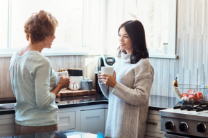Two women stand in the kitchen with cups of coffee, having hard conversations while caregiving.