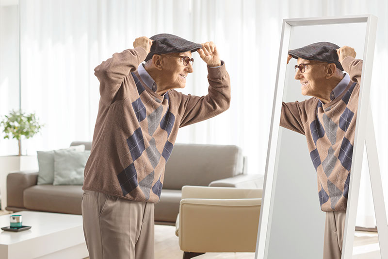An older man wearing the best senior adaptive clothing smiles at his reflection in the mirror.