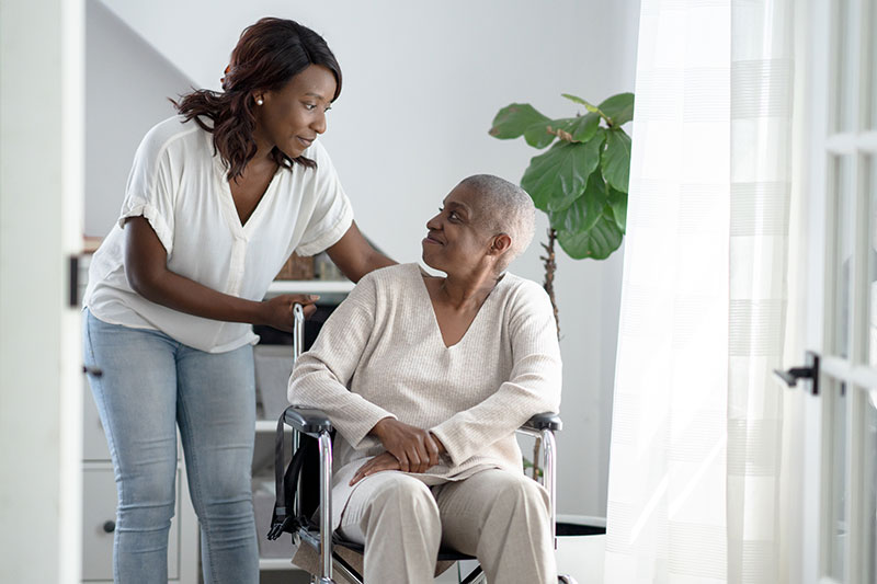 A woman in a wheelchair receiving hospital care at home is assisted by her adult daughter.