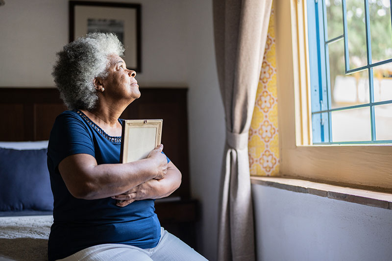 An older adult who is grieving clutches a photo in her arms as she gazes sadly out the window.