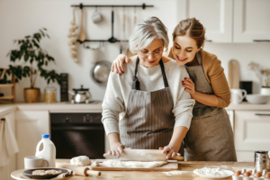 A young woman smiles over the shoulder of an older loved one as she rolls out biscuit dough, a great activity for someone with Alzheimer’s.