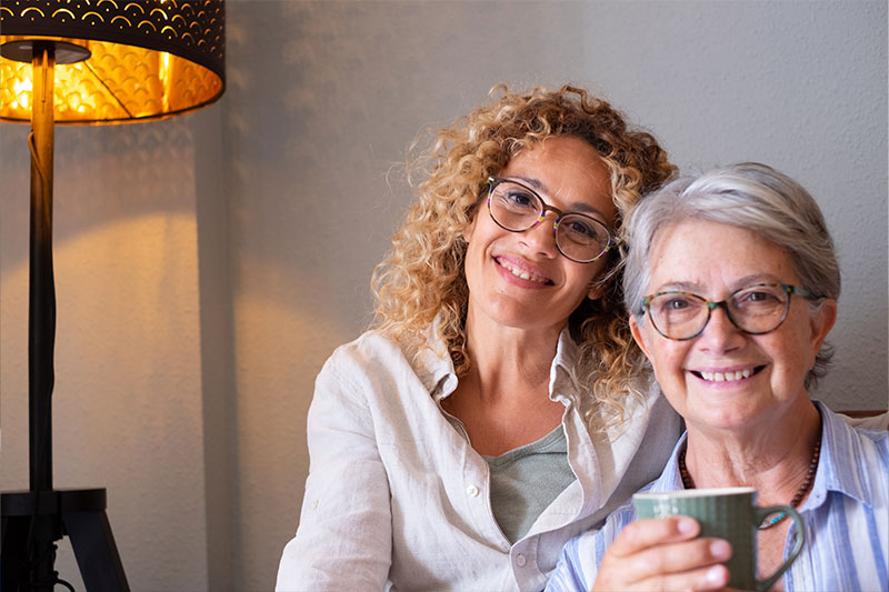 An older woman receiving 24-hour home care services in Fort Lauderdale smiles as she holds a cup of tea and sits beside her caregiver.