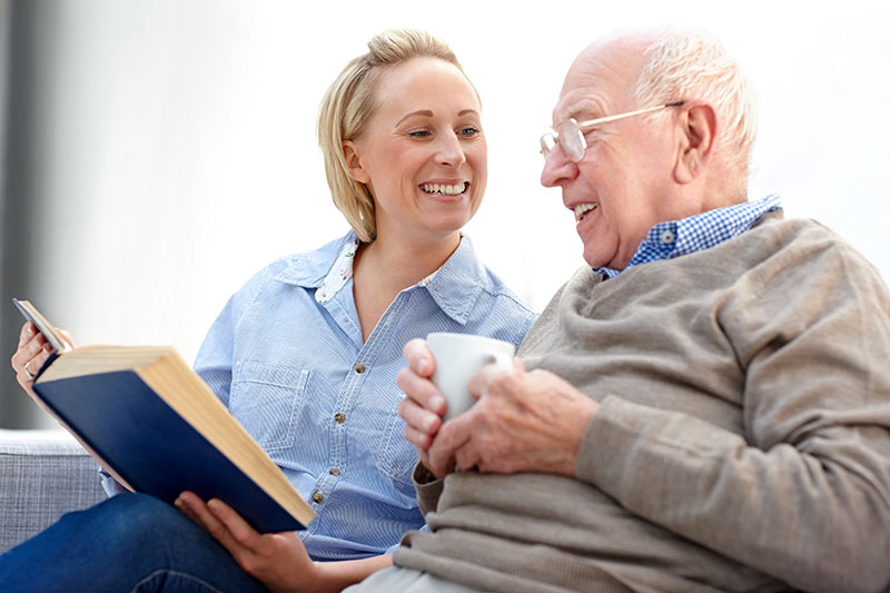 An older man receiving companionship services in Fort Lauderdale smiles as he enjoys a cup of coffee and the book his caregiver is showing him.