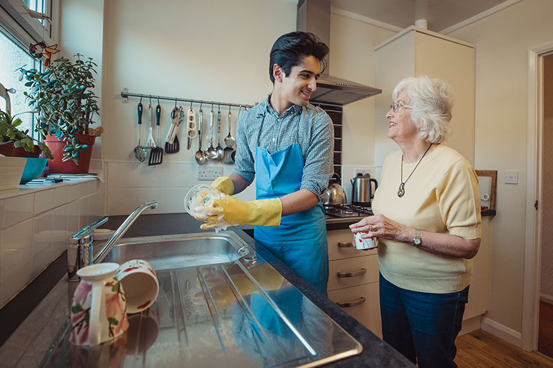 An older woman receiving light housekeeping services in Fort Lauderdale smiles at her caregiver as he washes dishes.