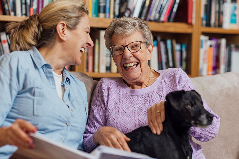 An older woman receiving memory care services in Fort Lauderdale laughs with her caregiver while holding her dog and looking at a photo album.