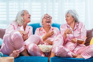 Three older women laugh as they’re aging well, sharing popcorn and a movie in their pajamas.