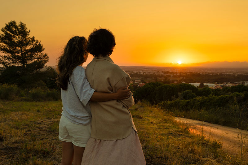 A woman who understands the link between medications and sundowning places her arm around her aging mother with dementia as they watch the sun set.